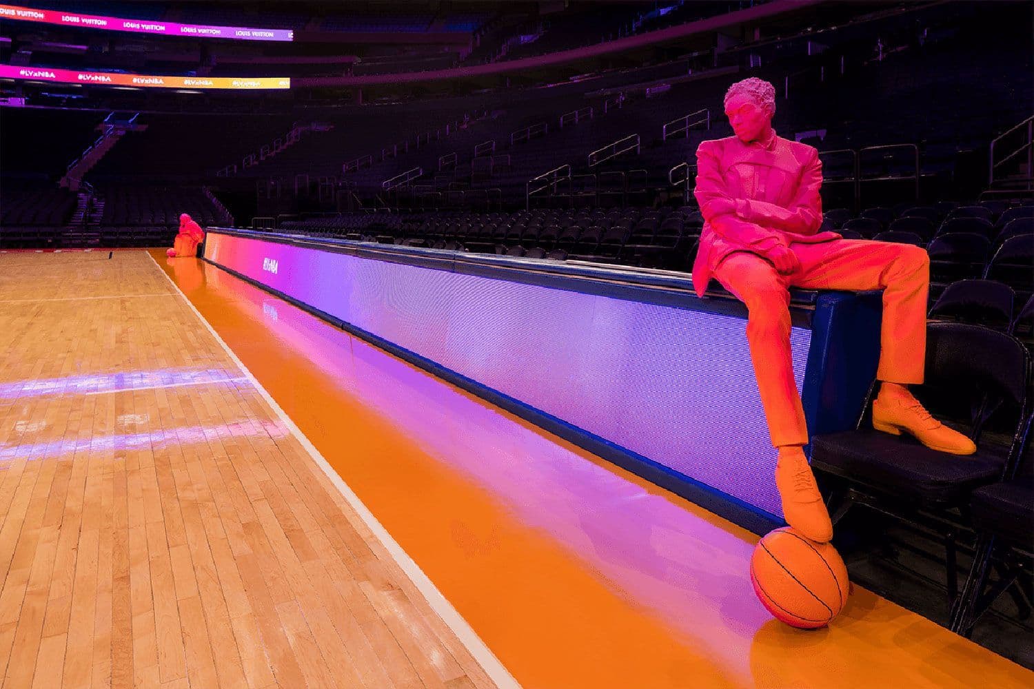Figure in a pink suit sitting on a courtside barrier rail beside a basketball on the arena floor.