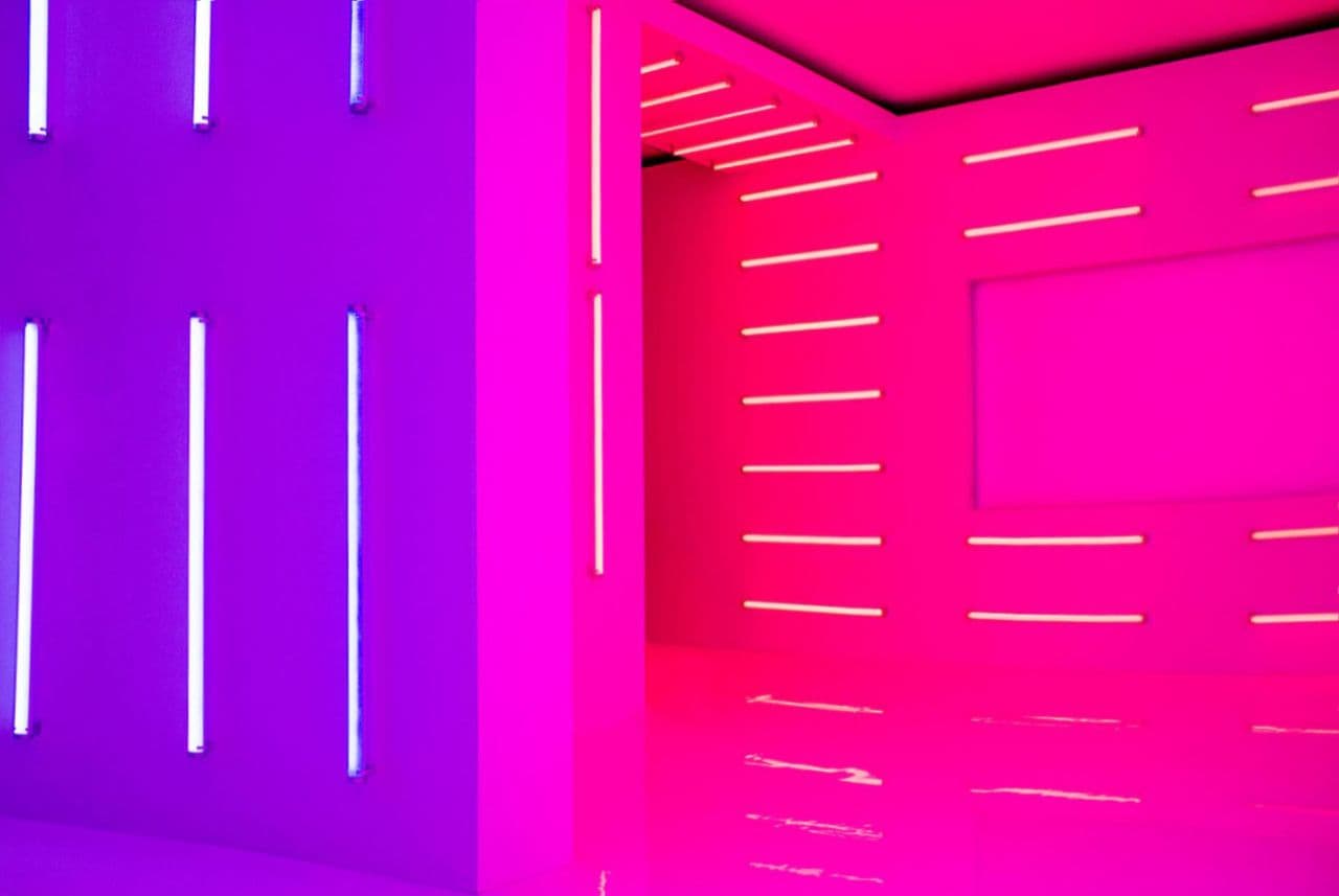 Neon-lit immersive room in purple and magenta, walls lined with glowing LED tubes reflected on a mirror floor.