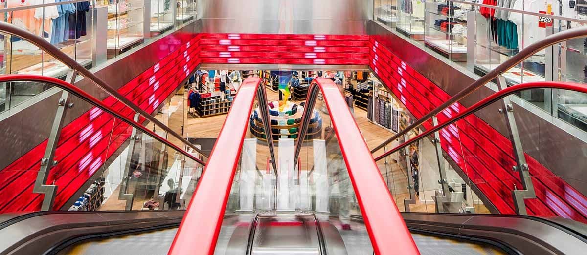 Mall escalator with red side panels leading down past a storefront with visible signage.