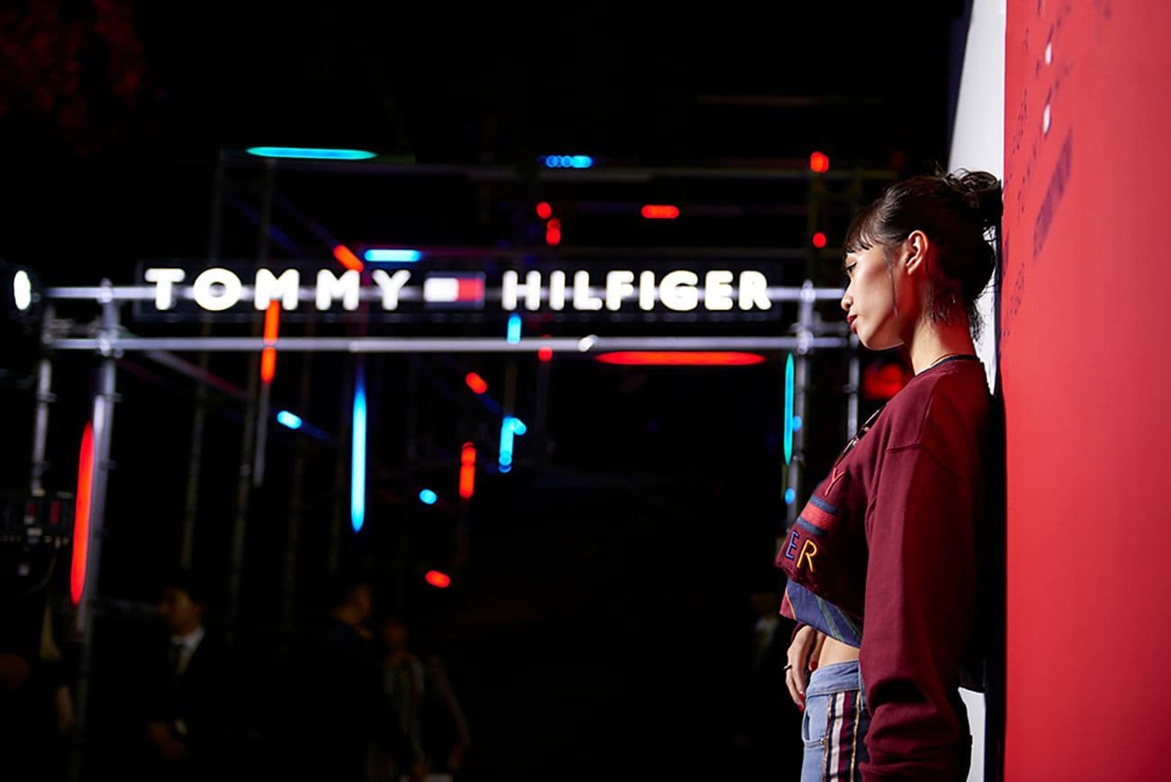 Woman standing beside an illuminated Tommy Hilfiger sign at night, with red and blue neon lighting in the background.