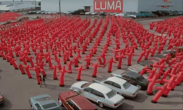 AI-generated video still of hundreds of red inflatable figures arranged in a grid across a car park, a 'LUMA' sign in the background.