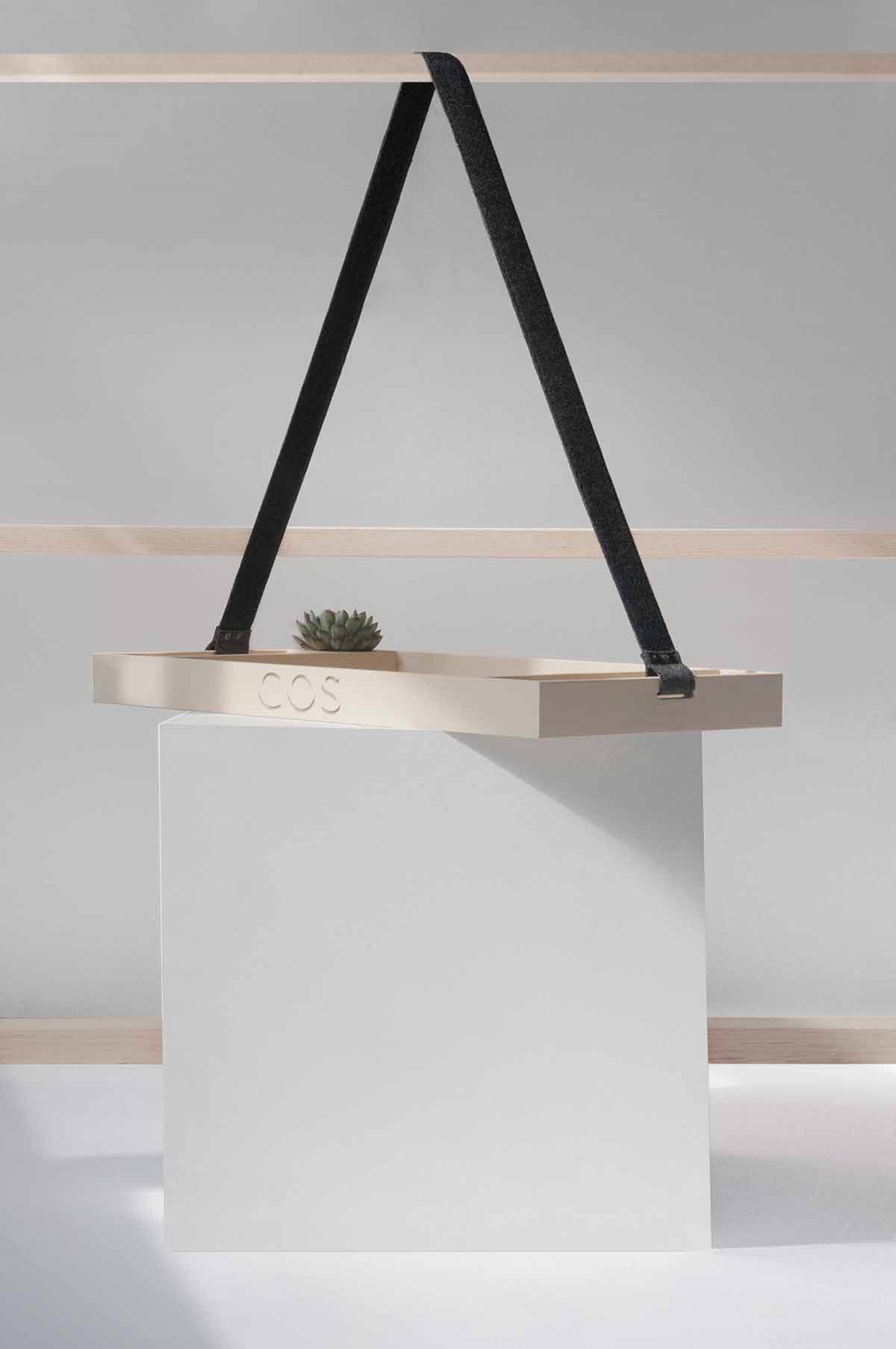 Plywood tray with COS branding suspended by a black strap over a white cube pedestal, styled with a succulent.