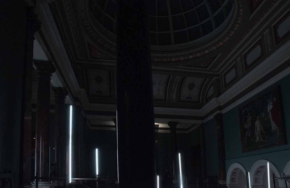 Wide shot of a domed museum hall with columns and vertical LED light strips, blurred figures, and a painting on the wall.