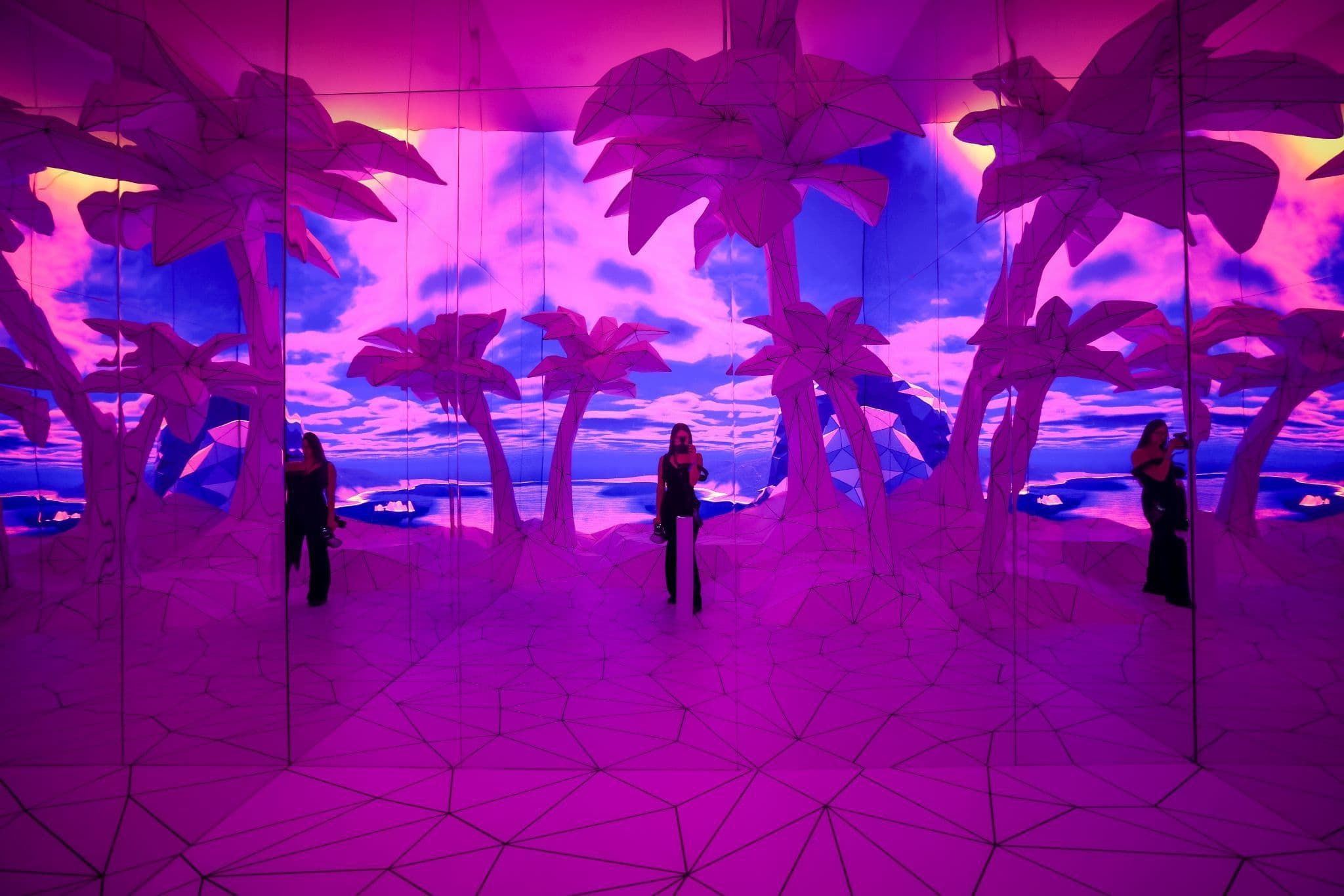 Mirrored room with pink and purple projected palm-tree scenery and two silhouetted visitors standing inside.