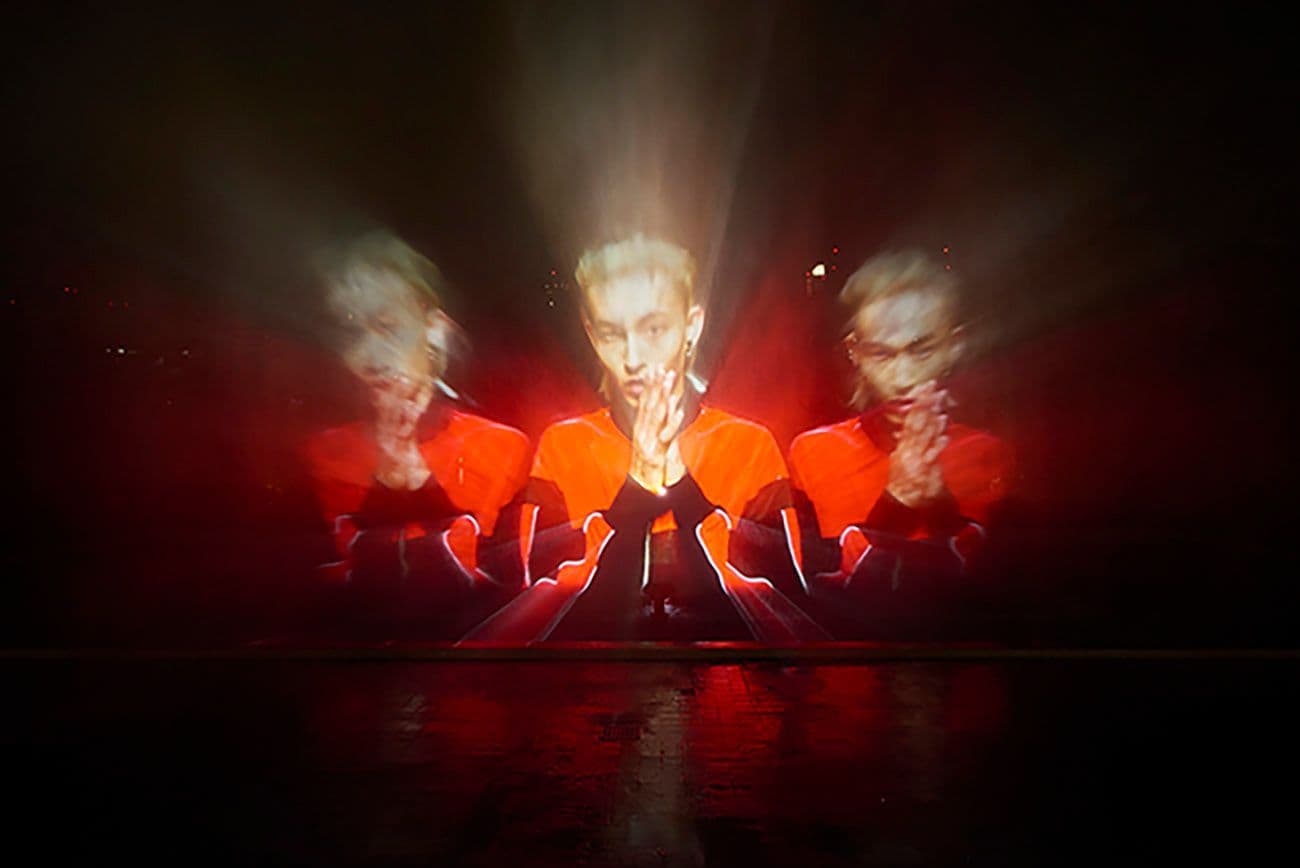 Person with hands raised in prayer pose, illuminated in red light with a mirrored double-exposure effect.