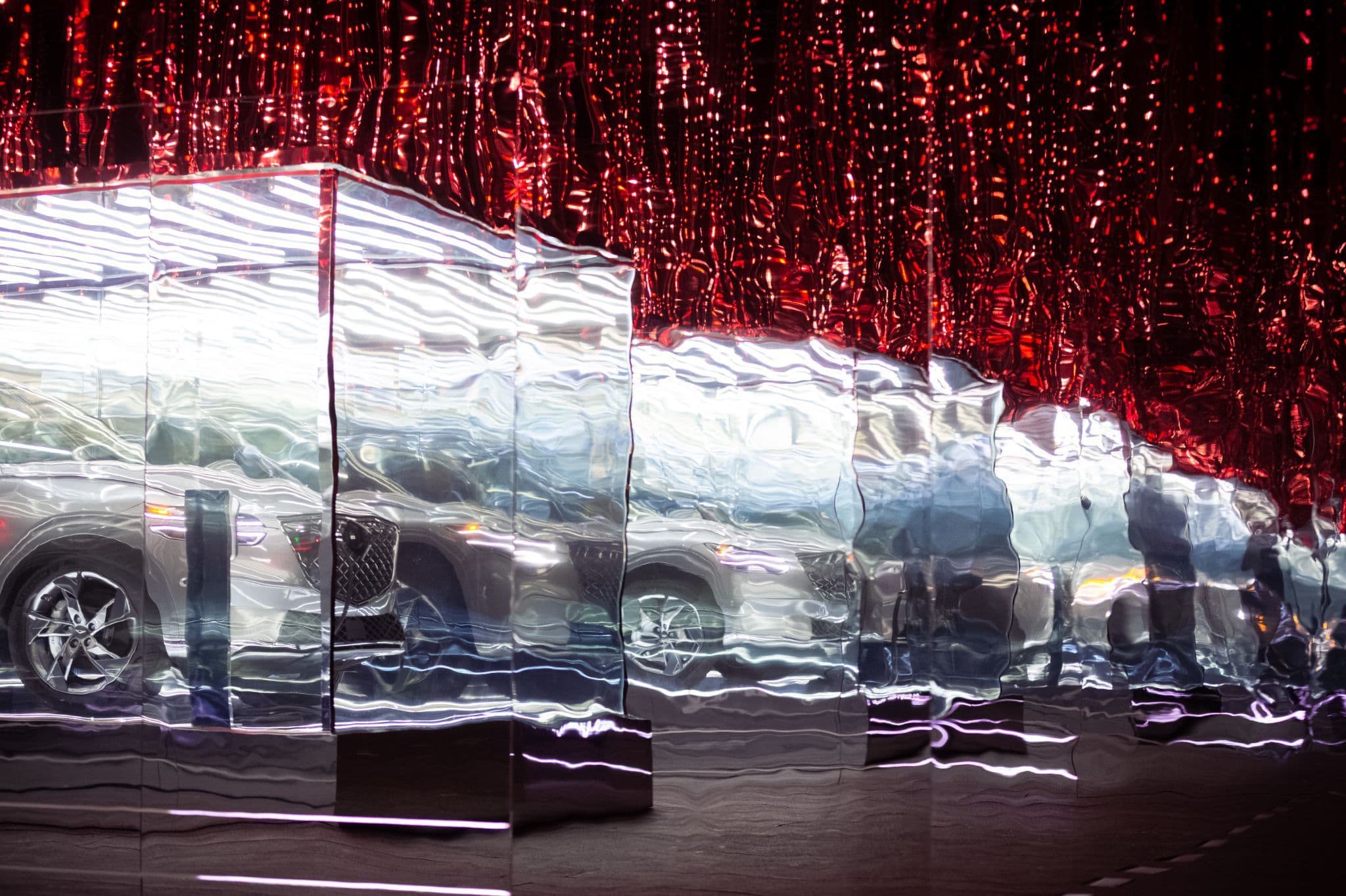 A car repeated diagonally across faceted mirror panels with a hanging red string-light curtain behind it.