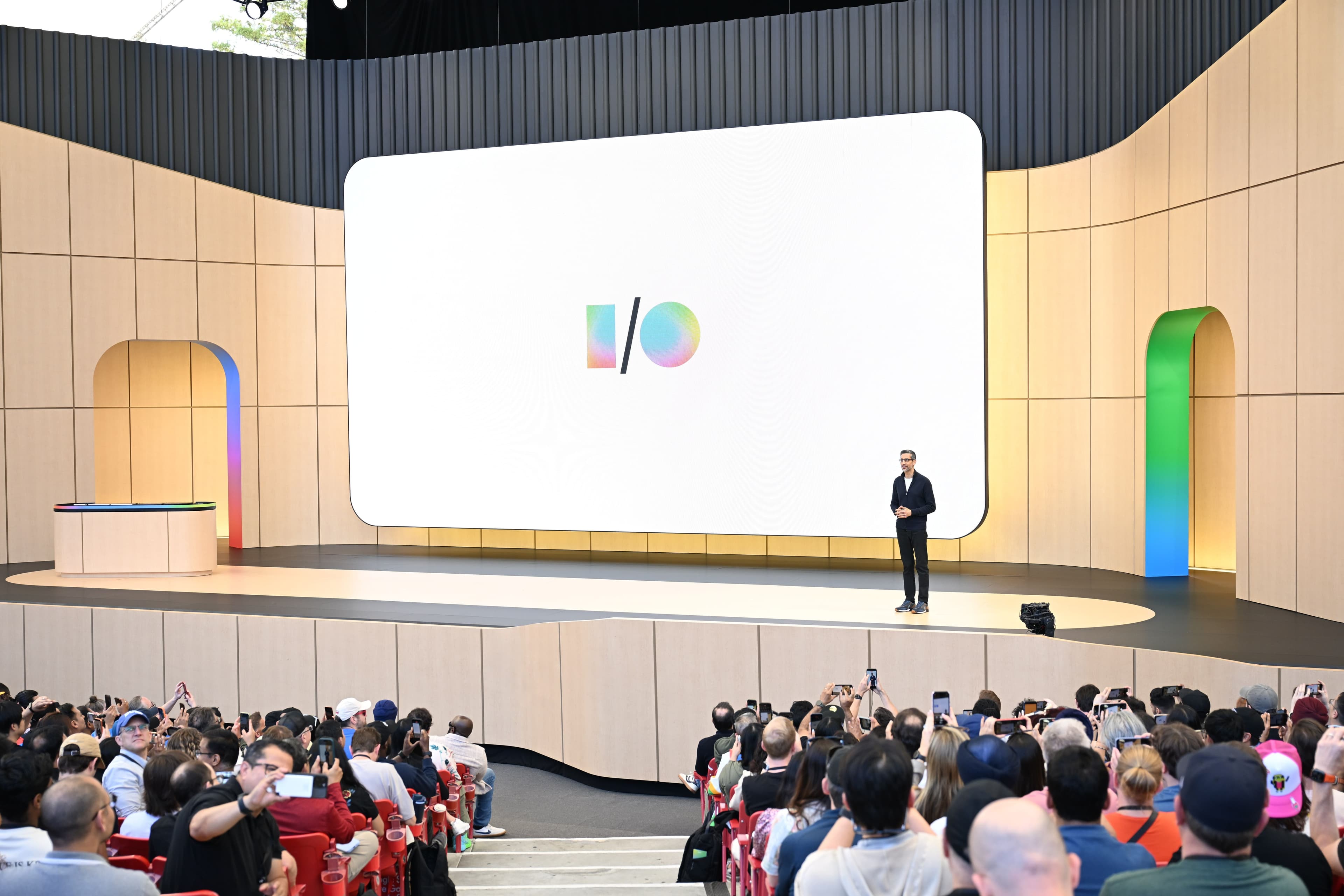 Presenter on stage beside a large screen showing the I/O logo, seated audience in the foreground.