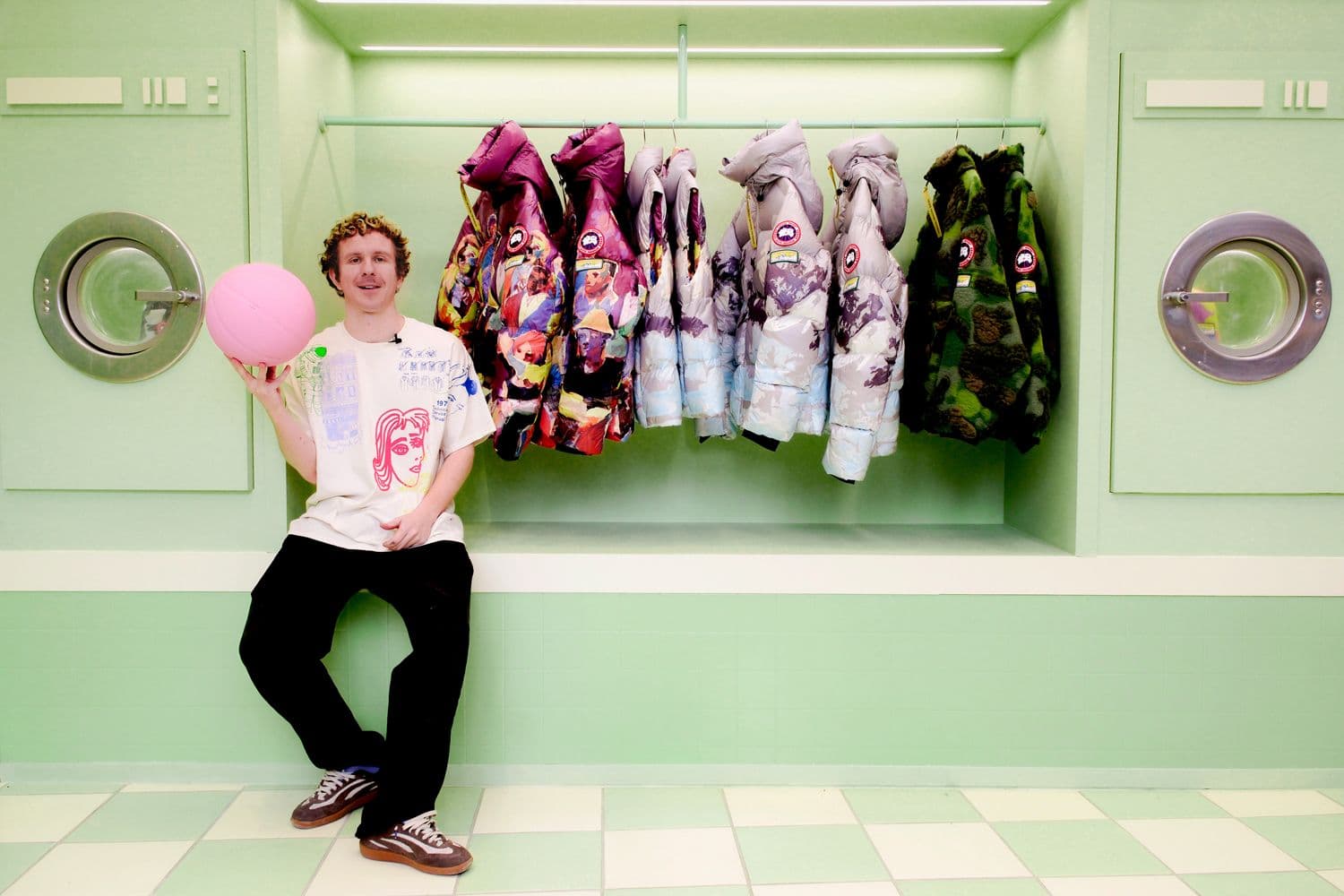 Man leaning against a mint-green wall holding a pink balloon, with printed hoodies hanging beside washing machine units