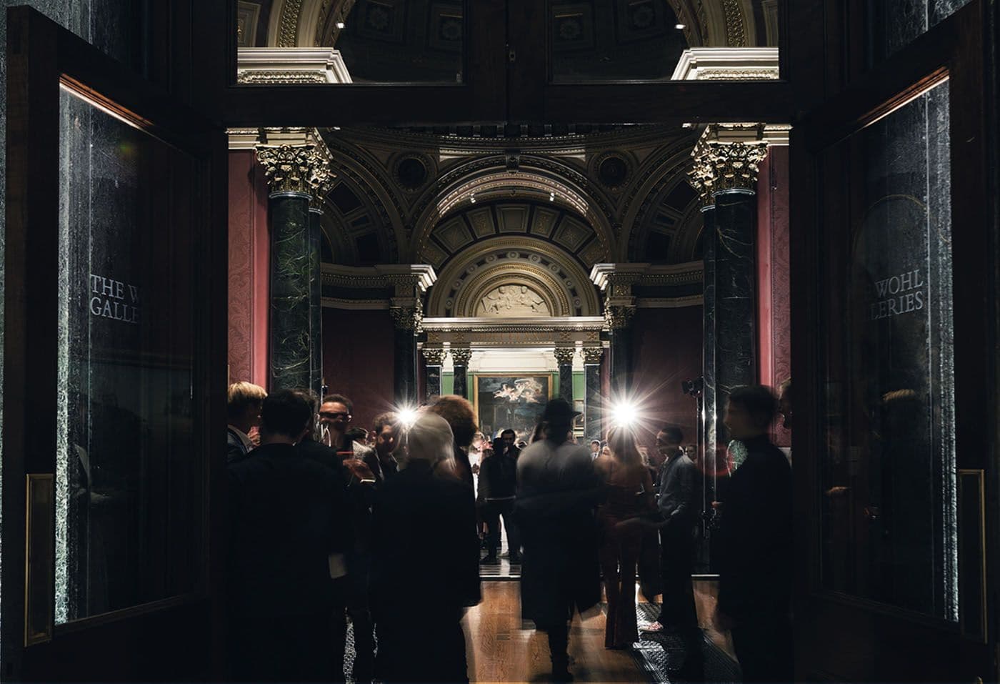 Crowd of guests silhouetted in a grand museum entrance hall with cameras flashing beneath an ornate archway.