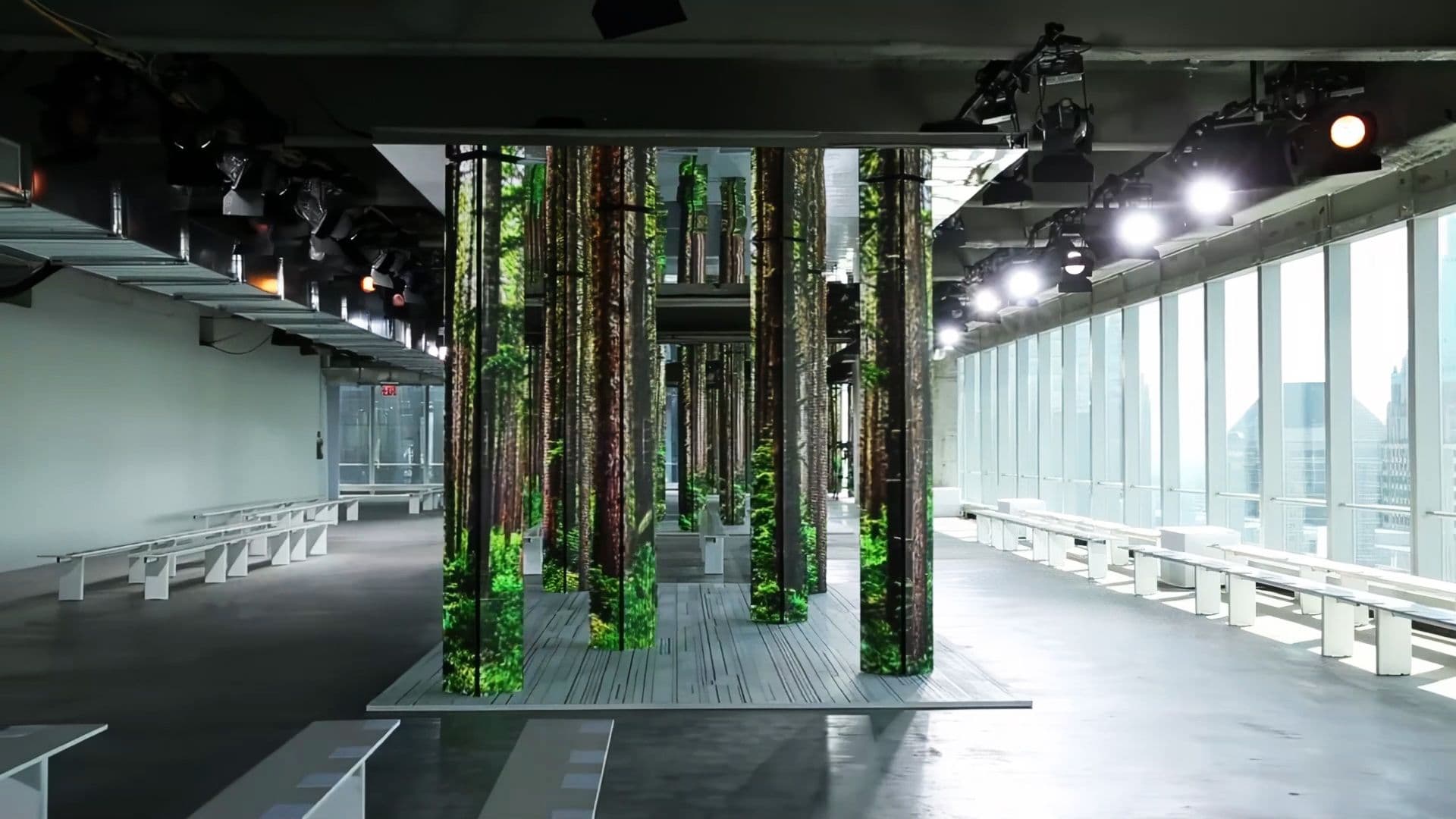Empty runway space lined with forest-print LED columns and rows of benches, city skyline visible through the windows.