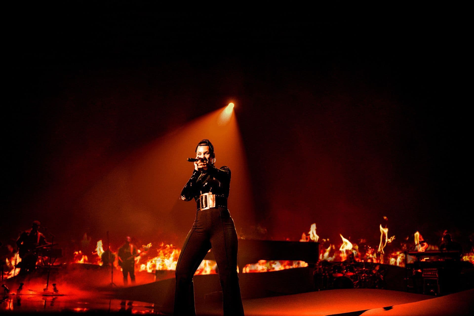 Performer standing under a spotlight singing on a dark stage with flame effects and band members in the background.