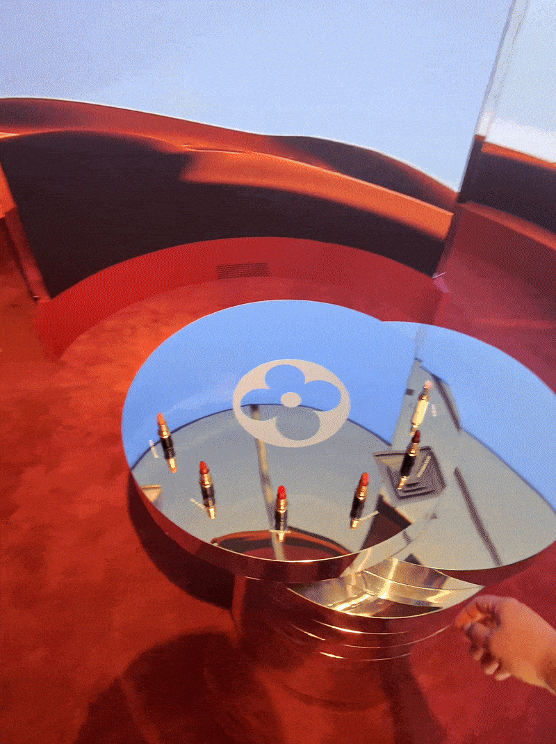 Overhead view of a hand reaching toward a round mirrored table etched with the Louis Vuitton flower monogram, ringed with lipsticks.