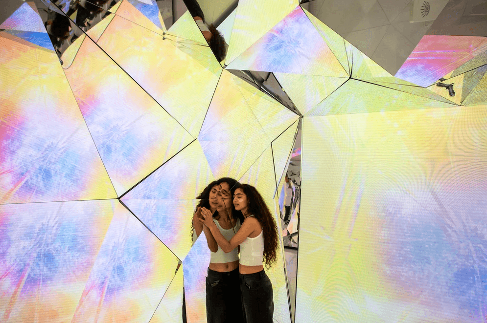Two women pose together inside a mirrored room paneled with colorful triangular yellow and pink glass.