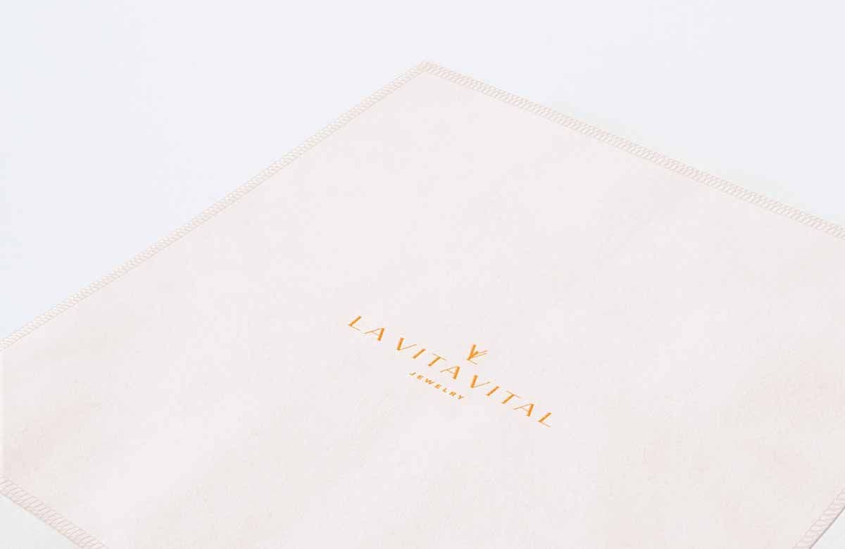 Cream cloth jewelry pouch with the La Vita Vital logo and wordmark printed in orange foil, laid flat and folded into a triangular shape.
