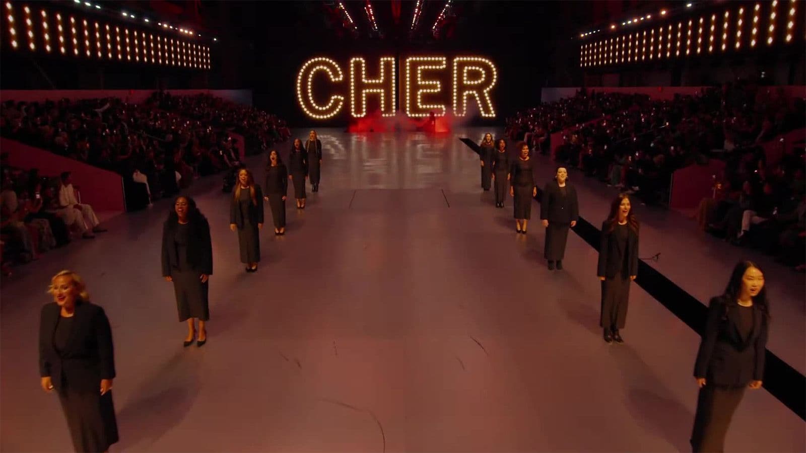 An illuminated "CHER" marquee sign glows above an empty runway lined with rows of seated audience members.