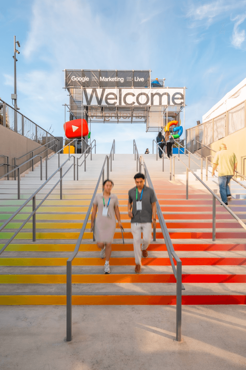 Rainbow-striped staircase leading up to a "Welcome" sign with the YouTube logo, two attendees walking up.