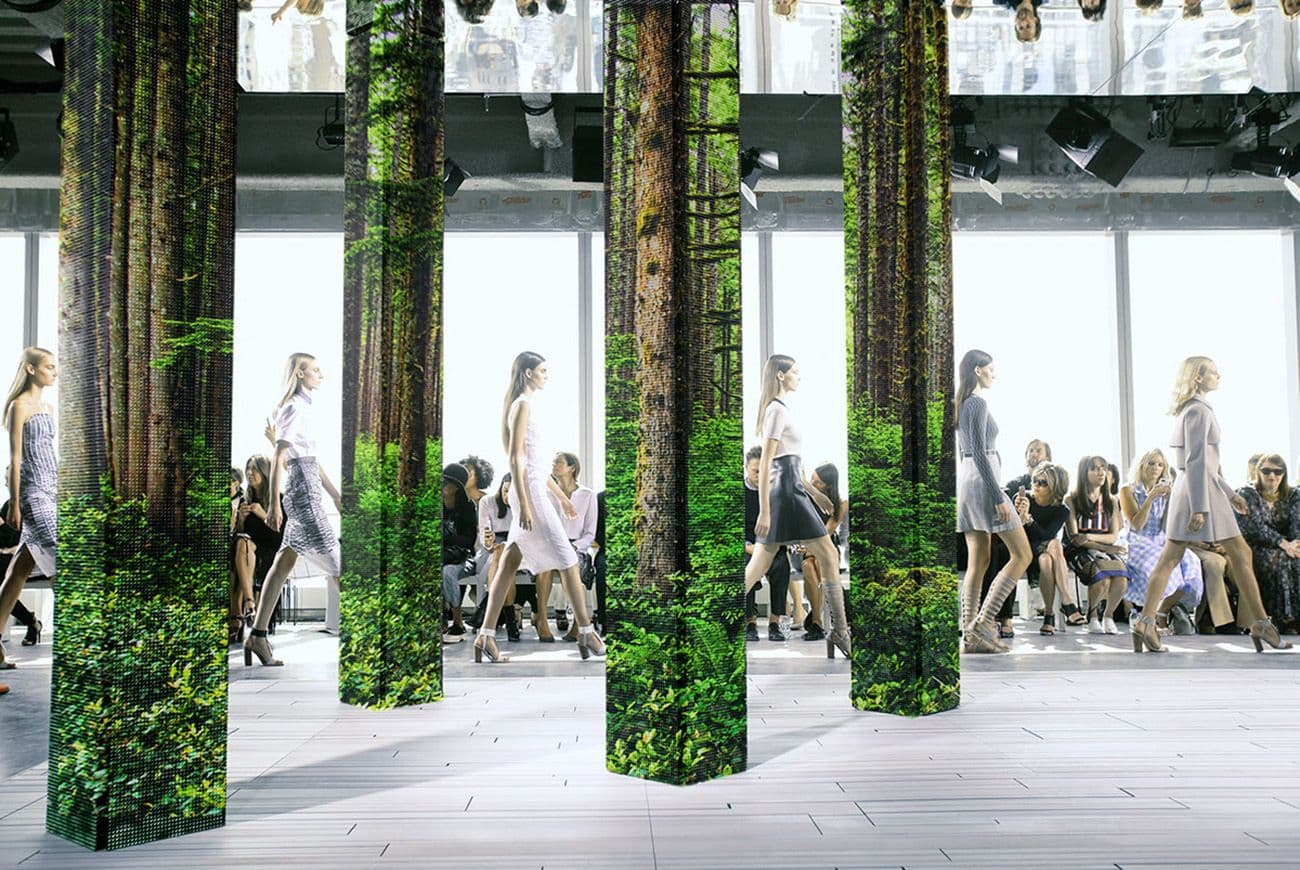 Wide view of a runway show with multiple models walking among tall columns wrapped in forest-print LED panels, seated audience on both sides, city skyline visible through floor-to-ceiling windows.