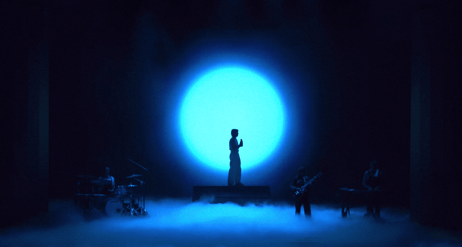 Silhouetted performer standing before a glowing blue circular light on a dark stage