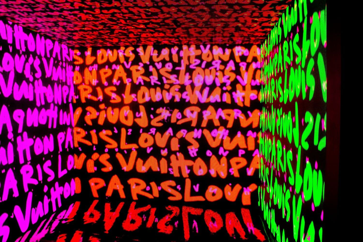 Cube-shaped installation with walls covered in overlapping neon Paris and Louis Vuitton script projections in pink, red, and green.