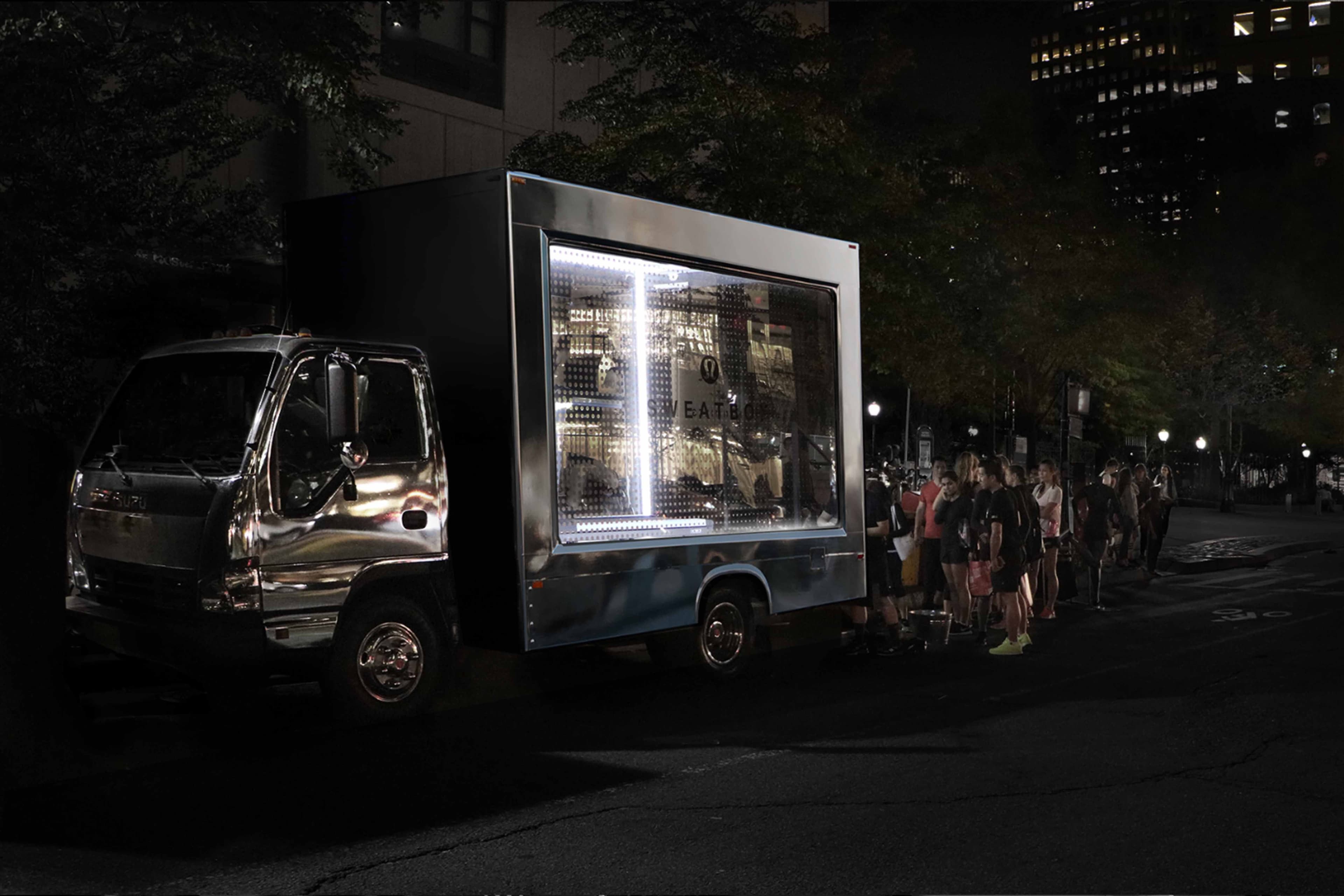 The mobile 'Sweatbox' truck parked on a city street at night with people lined up beside it