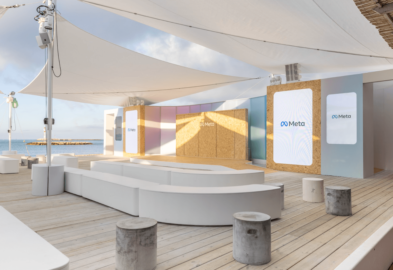An open-air lounge with curved white benches, pastel-colored wooden panels, Meta logo signage, and a view of the ocean beyond.
