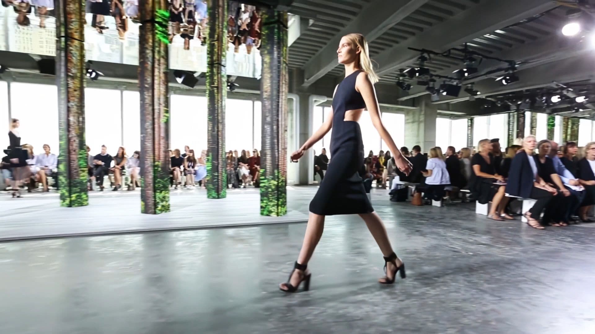 Model in a black dress and heels walking the runway among forest-print LED columns and seated audience.