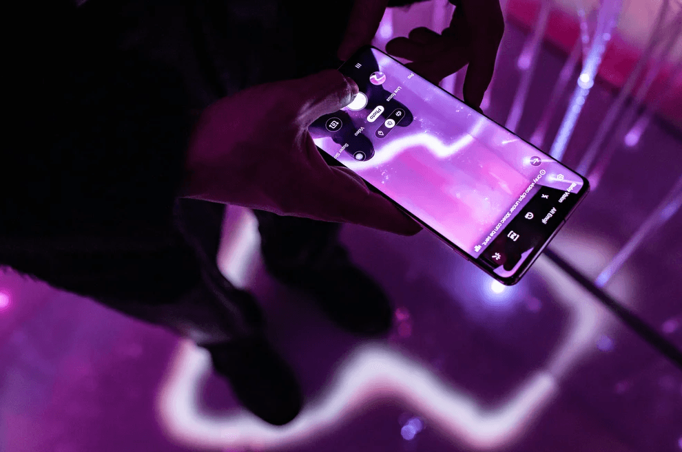 Close-up of a hand holding a smartphone with a squiggly line drawn on its screen against a purple fiber-optic backdrop.