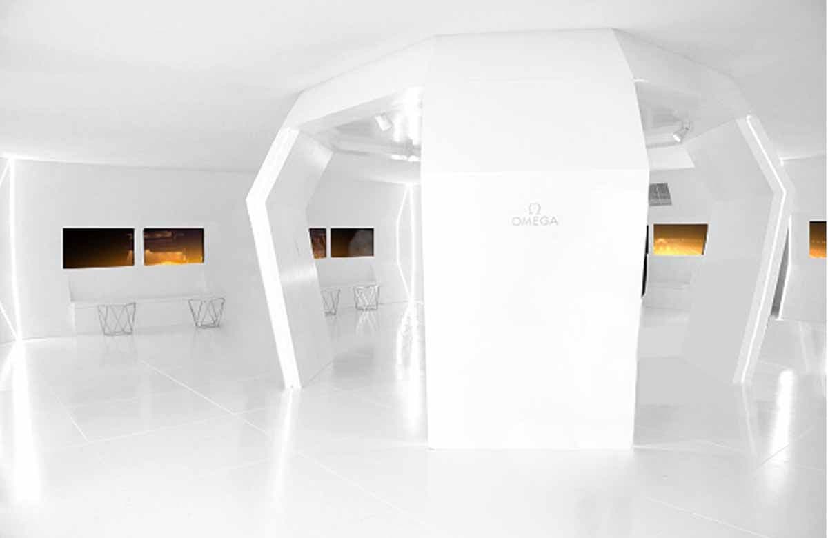White geometric exhibition hall with a central OMEGA-branded column and wall screens showing sunset and desert imagery.