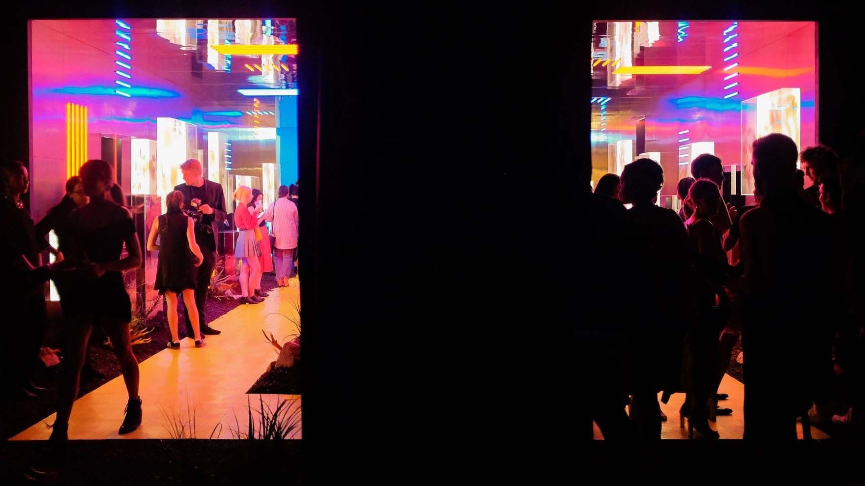 Two-panel view through dark doorways of a colorful mirrored room where guests mingle at an event.