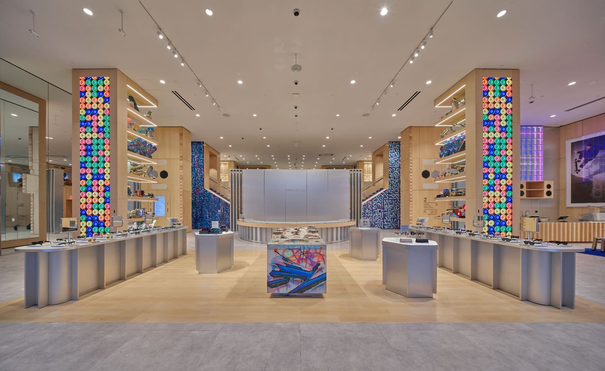 Open retail hall with light wood display towers featuring colorful patterned panels, a central staircase, and pedestal display tables.