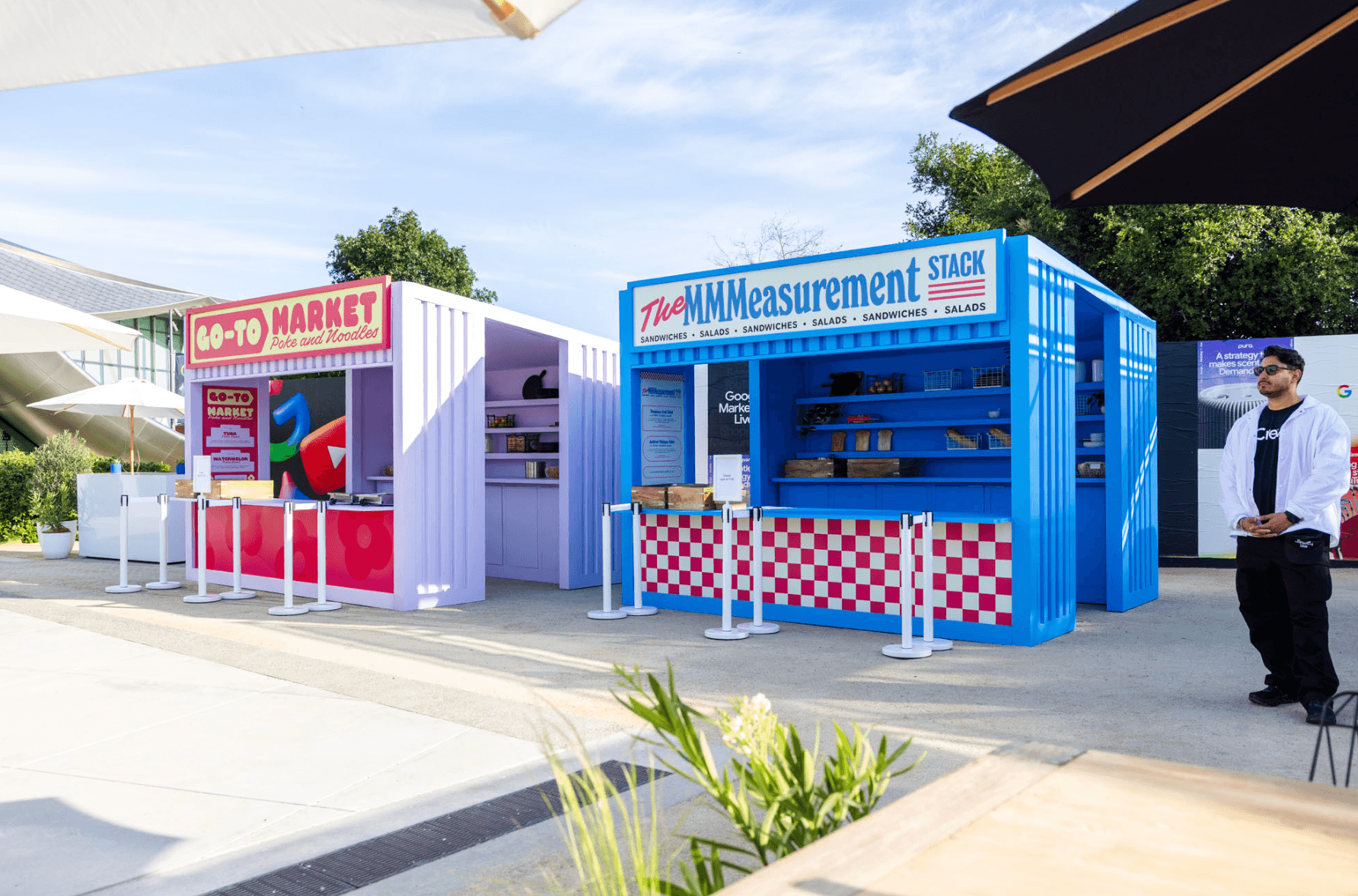 Row of colorful outdoor vendor booths with striped awnings, including a market-themed stall, under a shade structure.