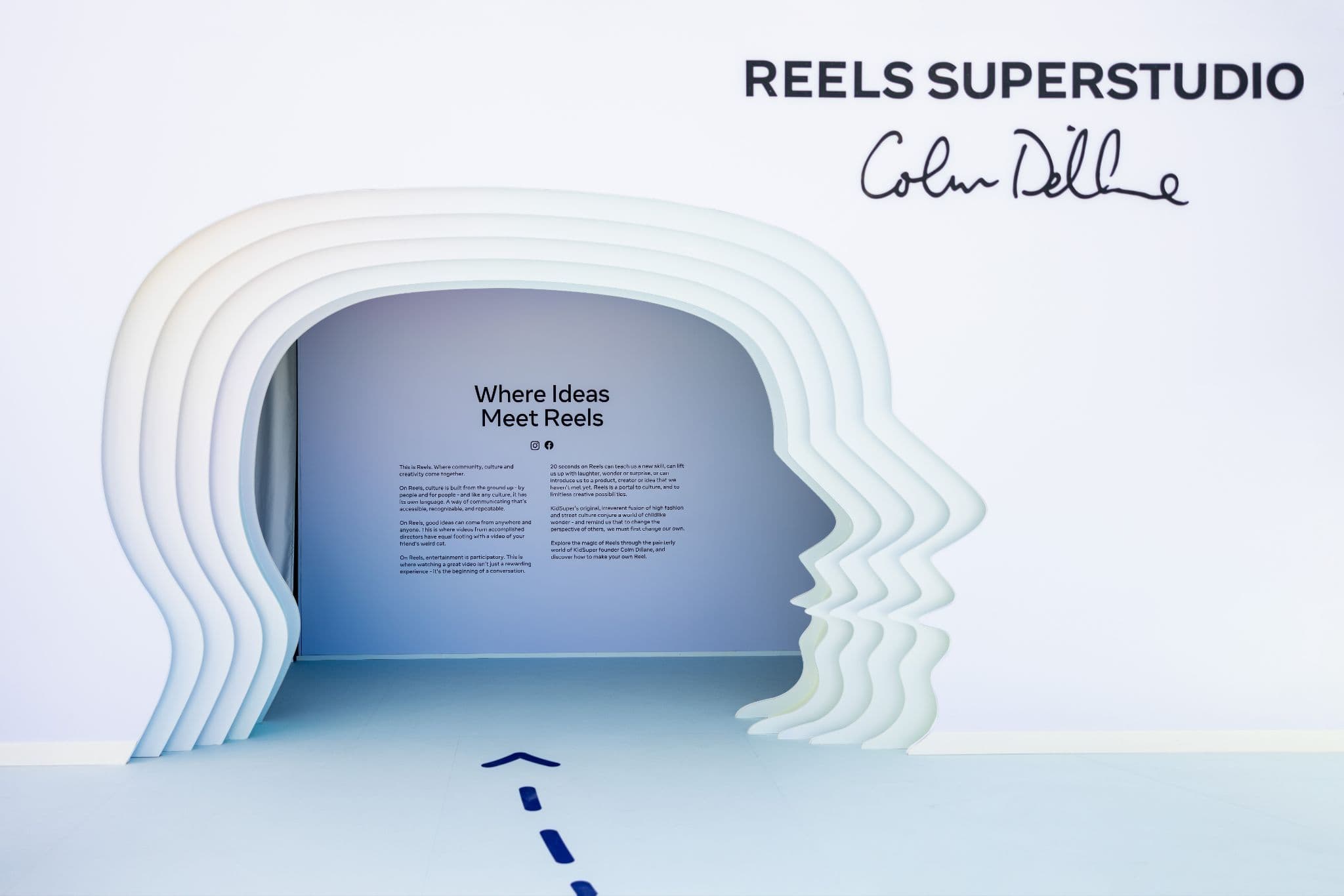 A white archway shaped like a stack of profile silhouettes leading to a gallery space with the text 'Where Ideas Meet Reels' and 'Reels Superstudio' signage above.