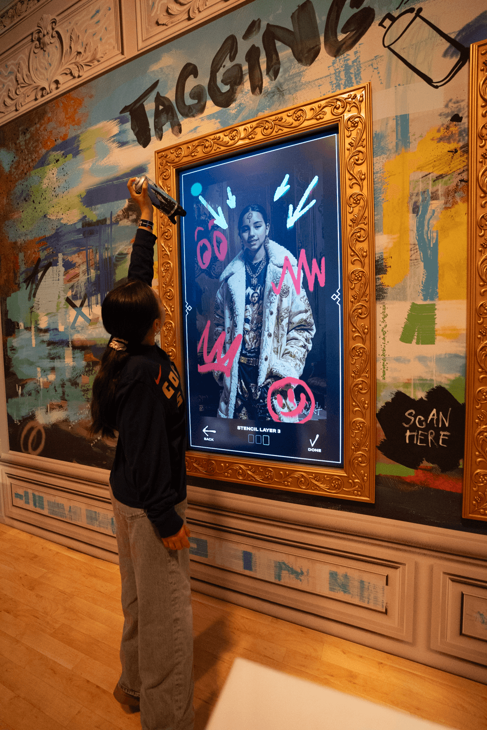 A person sprays a can of paint at a gold-framed digital screen showing a tagged portrait during an augmented graffiti activation.