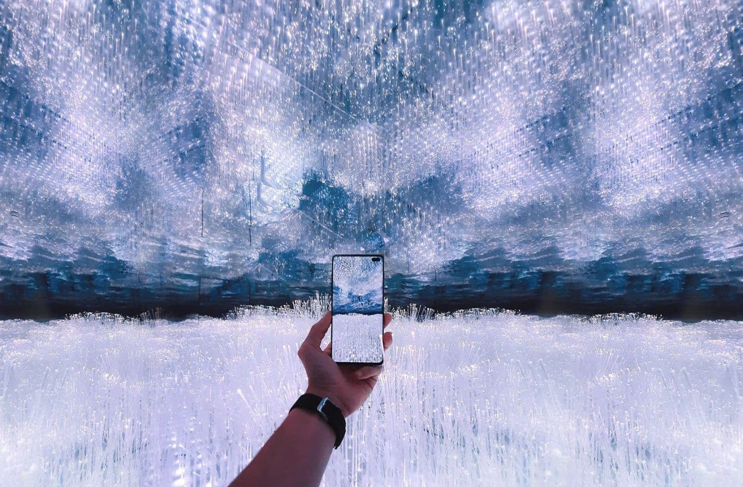 A hand holds up a smartphone against a mirrored room reflecting an overcast cloudy sky.