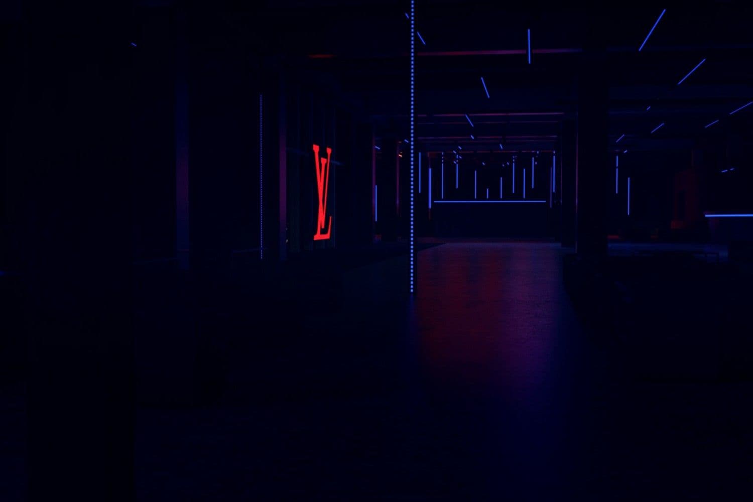 Dim exhibition corridor with a glowing red LV monogram sign and rows of ceiling lights receding into the distance.