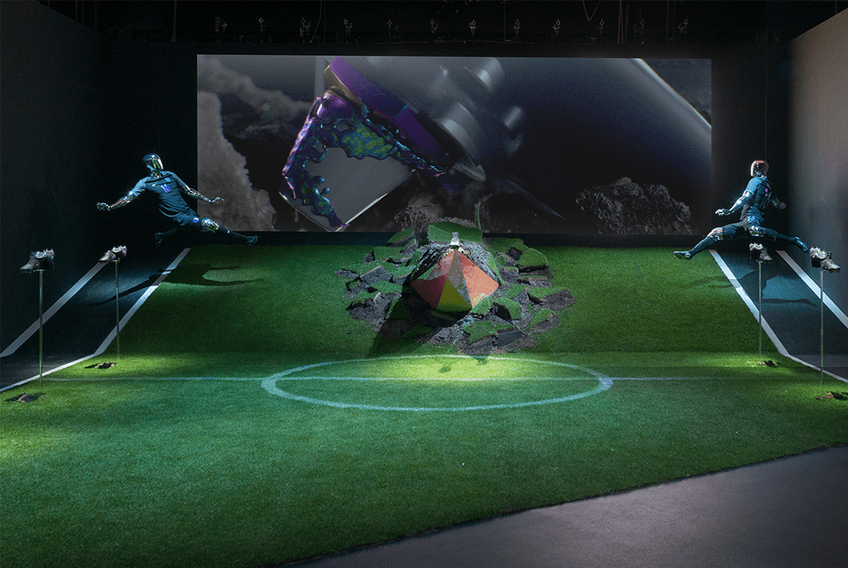 Installation on a green turf floor with two figures suspended on cable rigs facing a central pyramid-shaped object breaking apart, backed by a large screen showing abstract shapes