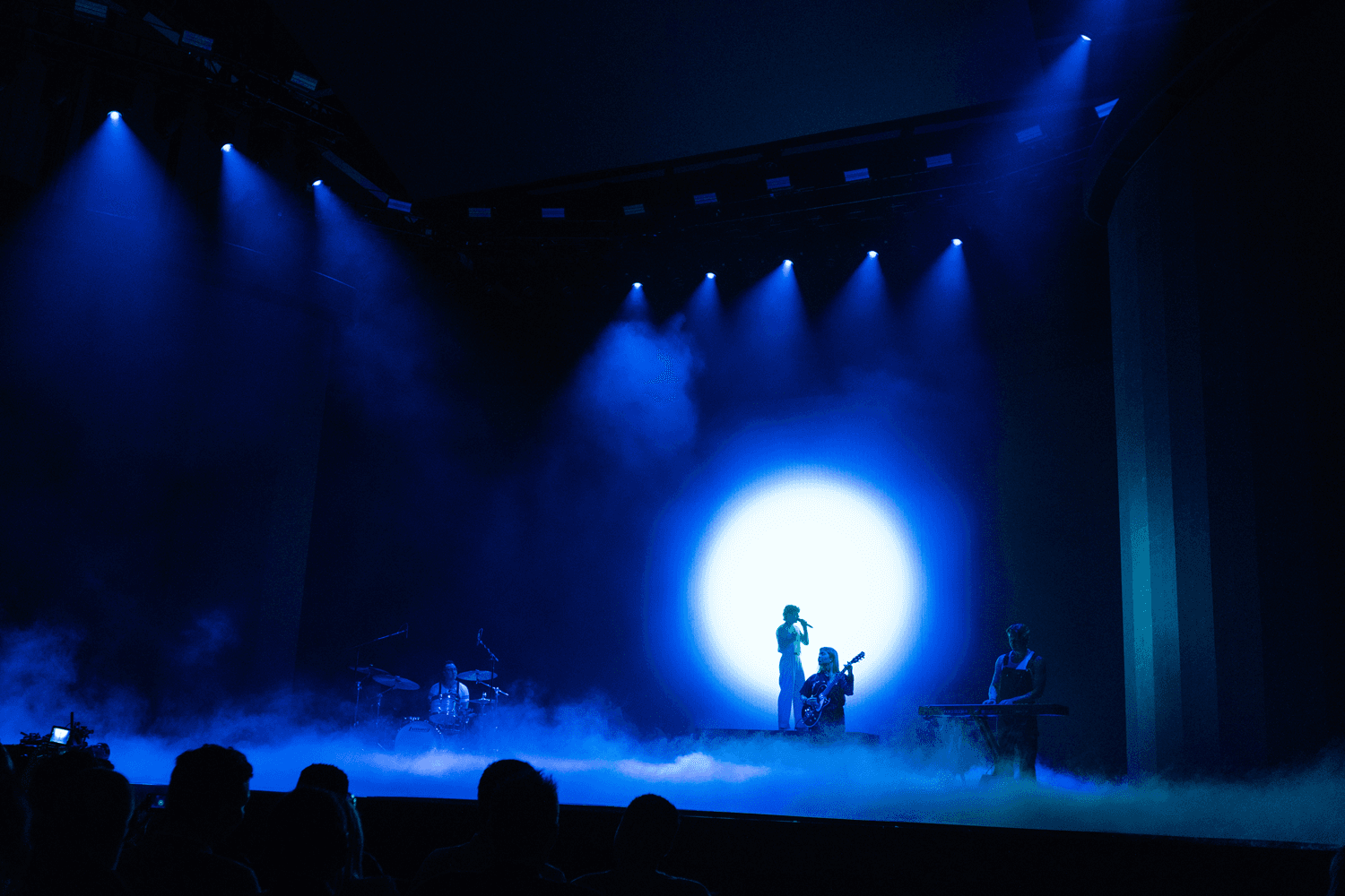 Silhouetted performer and musicians on stage beneath a glowing blue circular light