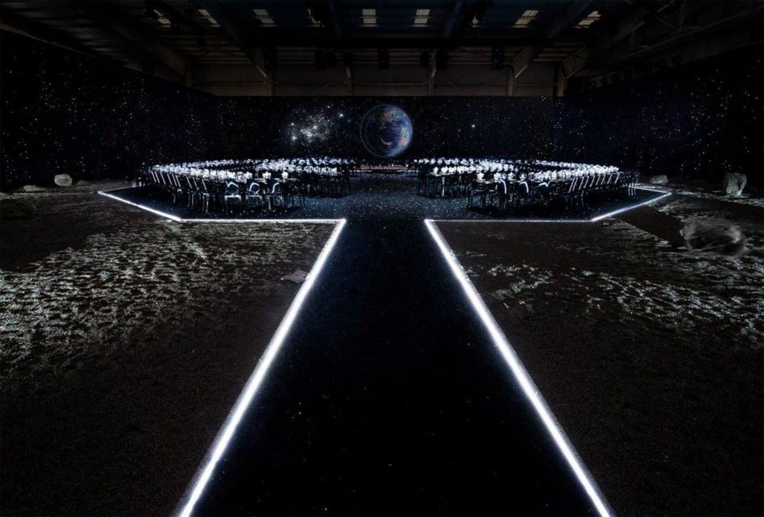 Wide hexagonal ring of dinner tables inside a dark hangar, with a glowing globe and starfield projected overhead.