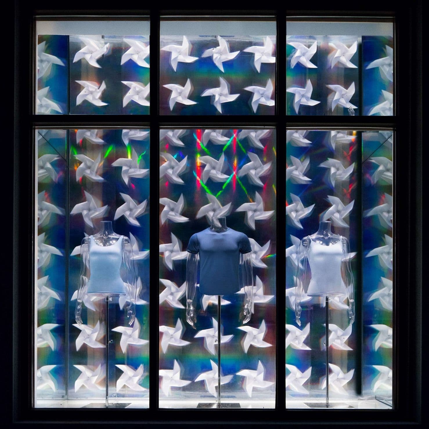 Storefront window filled with white pinwheel-shaped ornaments and rainbow light streaks over mannequins in shirts.