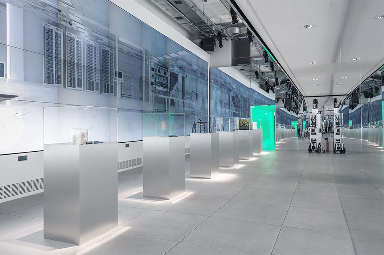 Exhibition hall with an illuminated video wall and a row of steel display pedestals down a corridor.