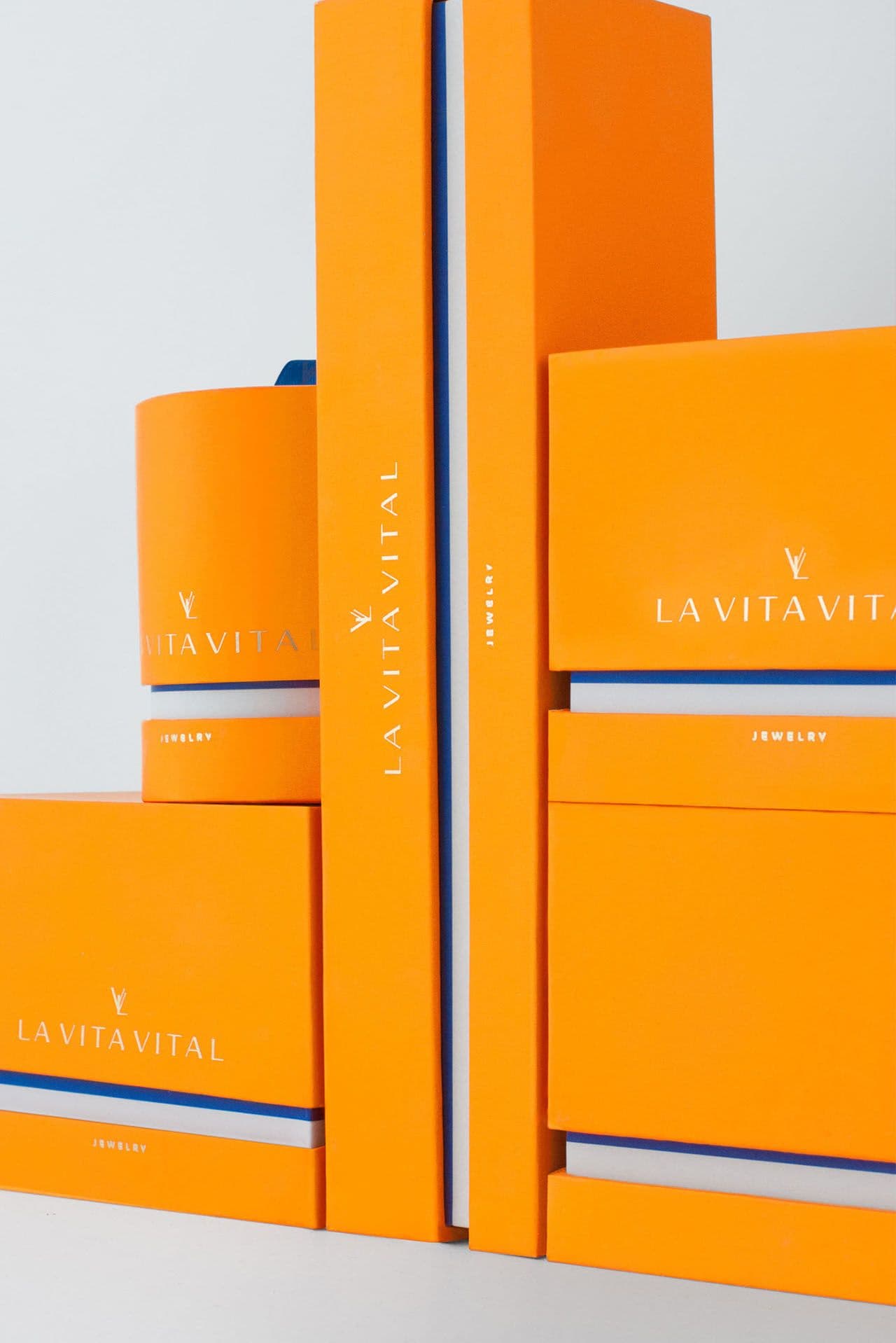 Tall vertical stack of orange La Vita Vital jewelry boxes with white logo and 'JEWELRY' text, separated by a thin blue and white striped book-style case.