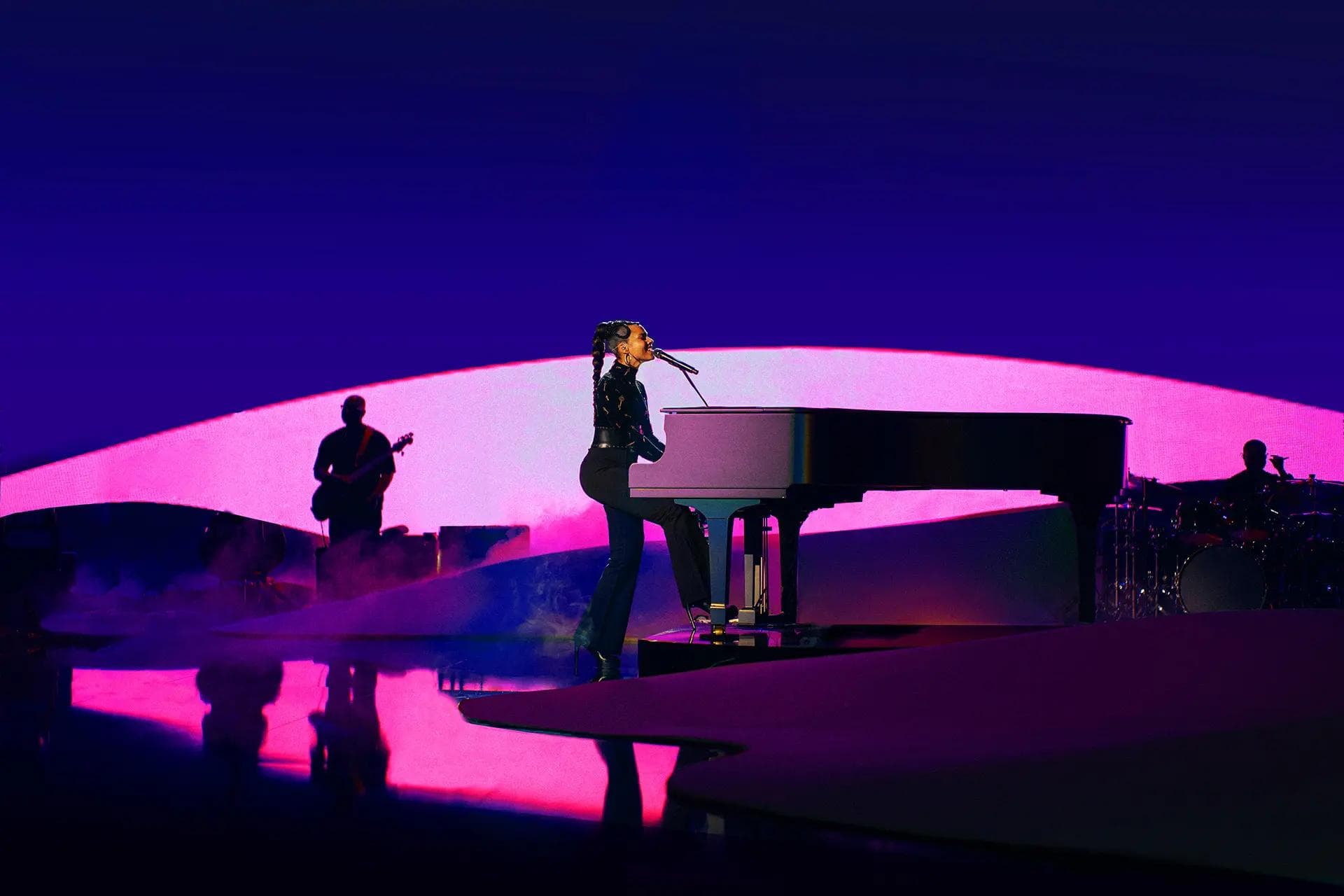 Silhouetted performer singing at a grand piano on a curved pink-lit dune stage, band members silhouetted behind against a deep purple backdrop.
