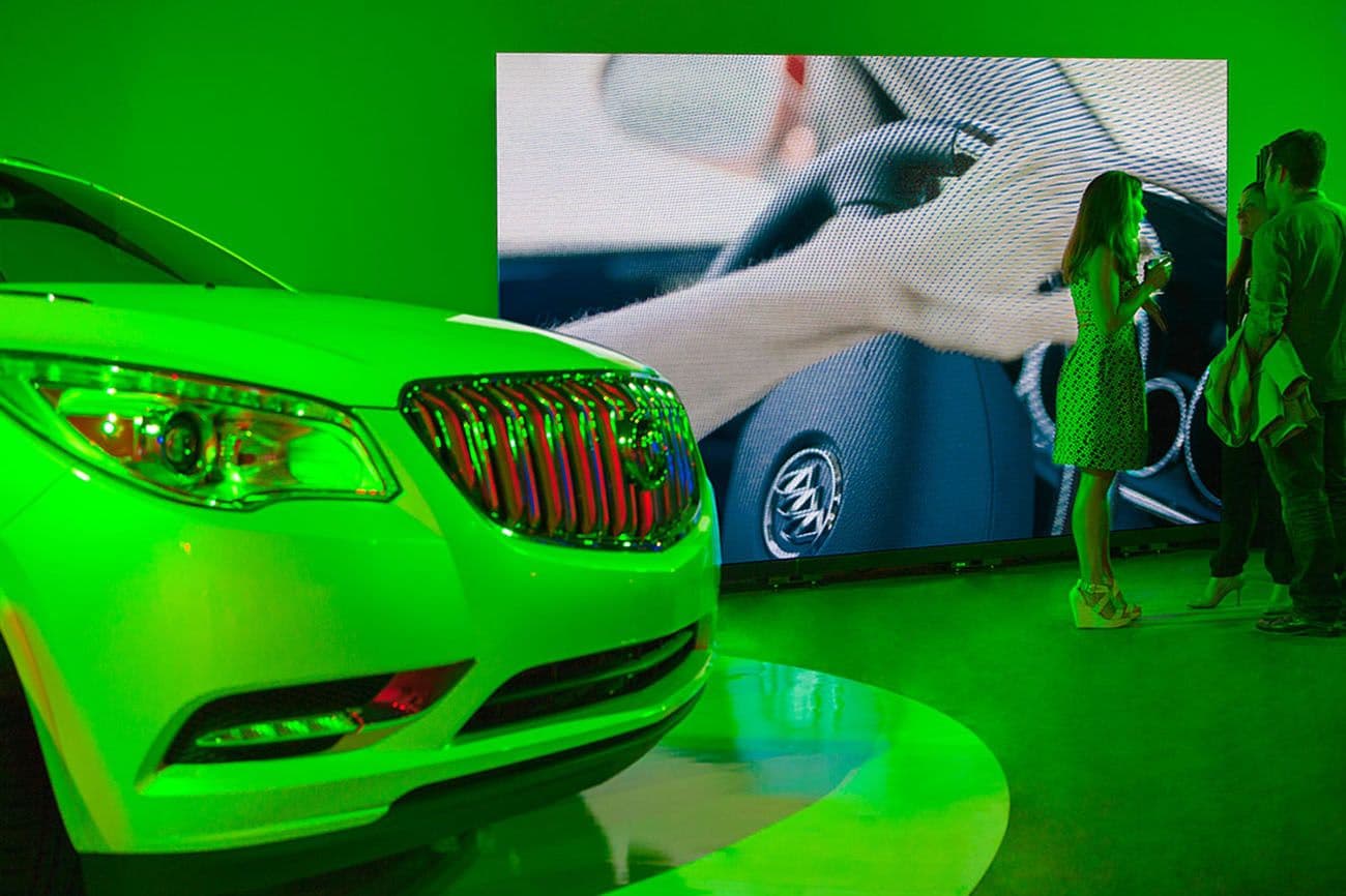 A green-lit Buick SUV on a display turntable in front of a video screen showing a hand on a steering wheel, with visitors watching nearby.
