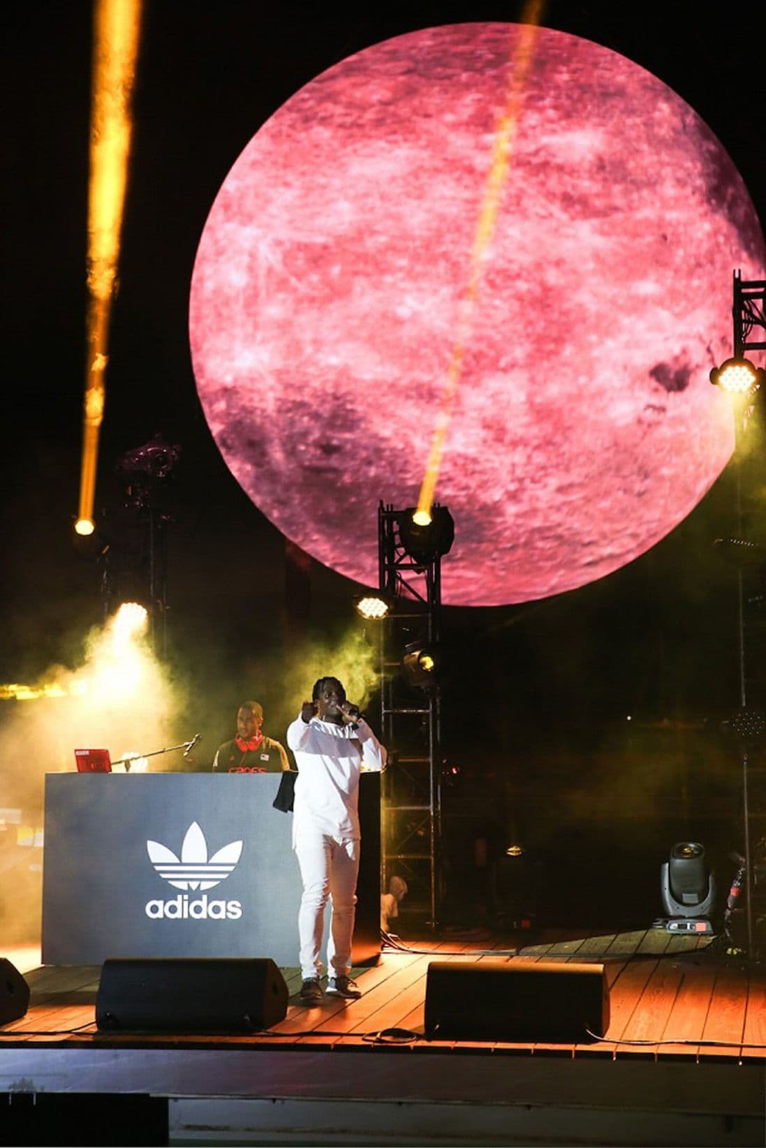 Performer in white singing into a microphone on stage in front of an adidas-branded DJ booth, with a large pink moon projection and spotlights overhead.