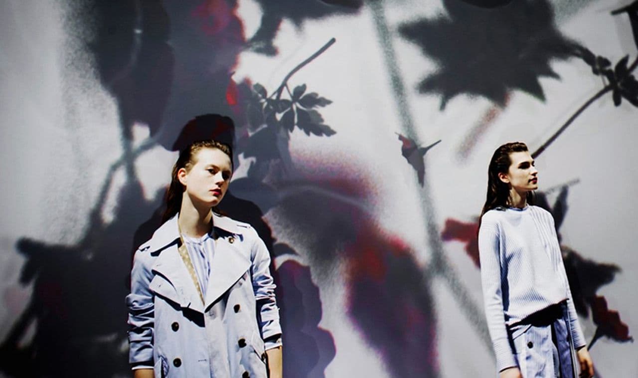 Two models in light-colored jackets standing before a wall projected with dark floral branch shadows.