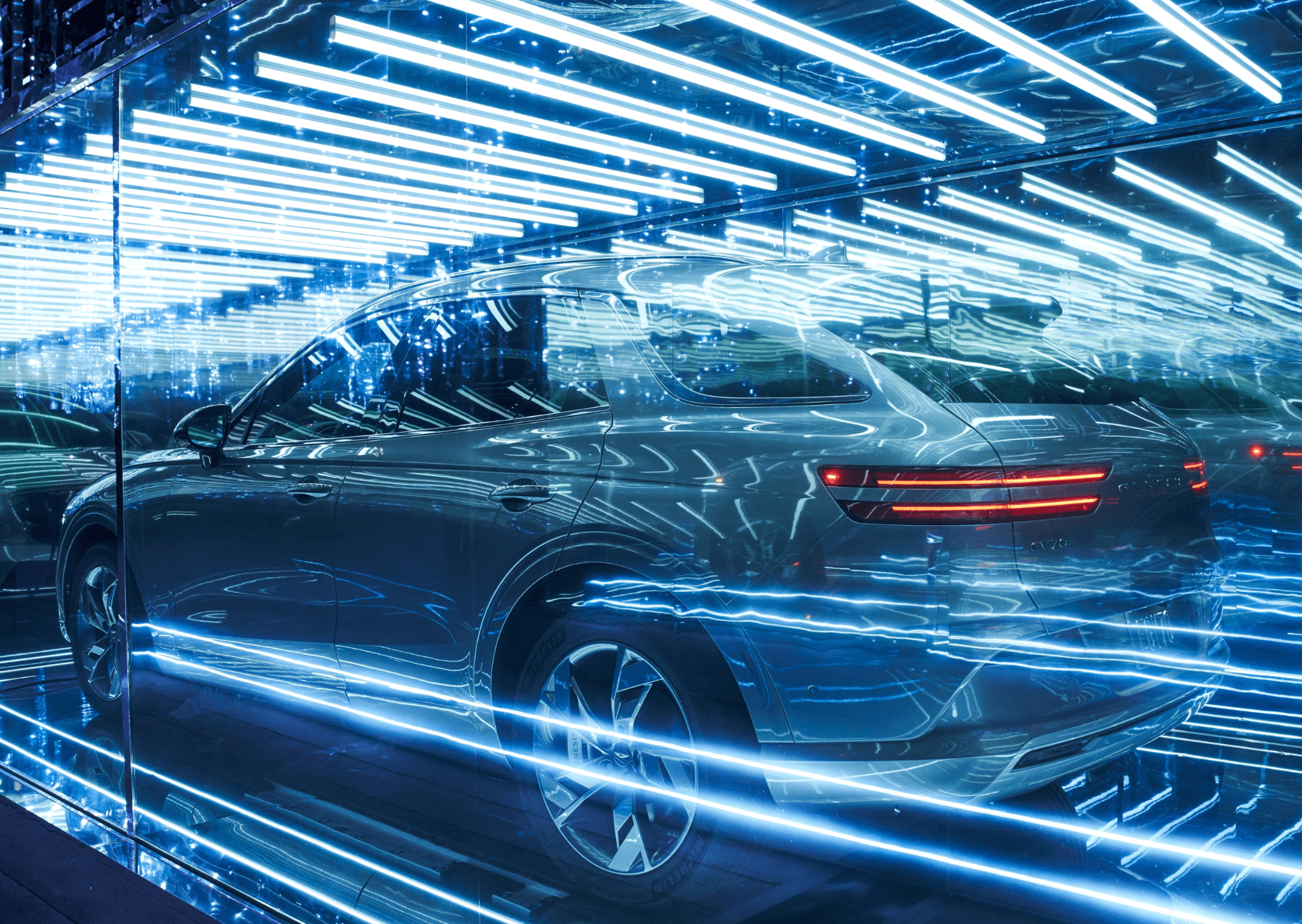 Side profile of a car reflected in mirrored panels under a ceiling of blue neon light tubes.