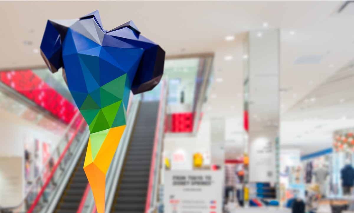 Multicolored faceted low-poly sculpture hanging in a bright mall corridor beside an escalator, storefronts in the background.