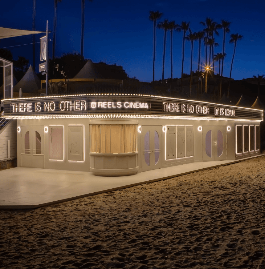 A beachfront theater building lit up at night with a marquee reading 'There is no other, Reels Cinema' and palm trees silhouetted against the sky.