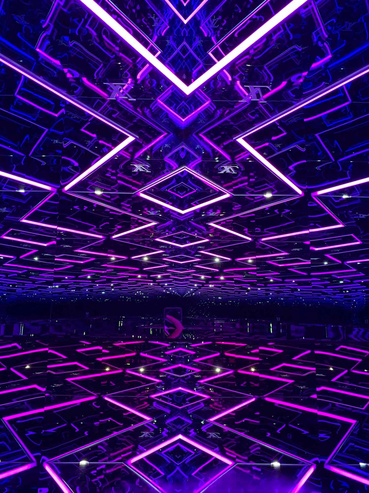 Purple neon X-shaped light installation reflected across a mirrored floor and ceiling.
