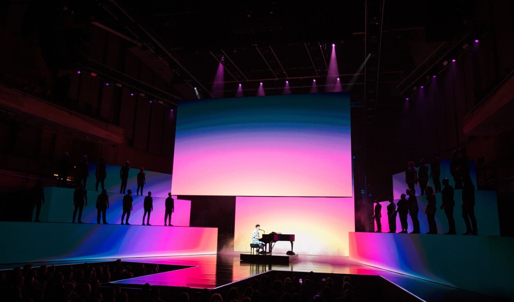 Performer at a piano on stage with a large pink and blue gradient screen behind, flanked by silhouetted dancers